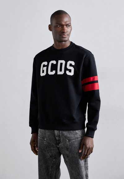 Sweatshirt noir avec logo "GCDS" blanc et accents rouges rayés sur les manches. Présente un col rond et des poignets côtelés. Associé à un denim gris clair.