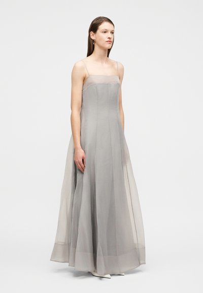 Γυναίκα που φοράει ένα μακρύ, ανοιχτό γκρι διάφανο φόρεμα με λεπτές τιράντες, στέκεται μπροστά σε απλό ανοιχτό φόντο, κοιτάζοντας ελαφρώς προς τα δεξιά.