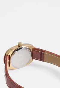 Gullfarget armbåndsur med brunt teksturert lærrem og rund kasse. Har en krone og en klar glassbakside.