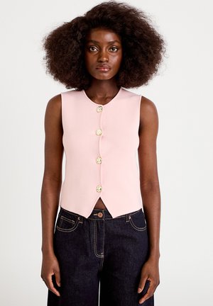 Woman with afro hairstyle wearing a sleeveless pale pink buttoned top and dark high-waisted jeans, standing against a white background.
