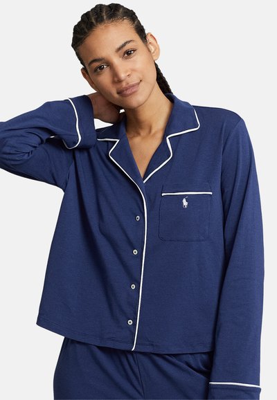 Cămașă de pijama bleumarin, confecționată dintr-un material moale, cu dungi albe, mâneci lungi și un buzunar pe piept cu un logo brodat.