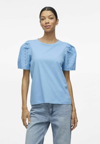 Jasnoniebieska bawełniana koszulka z krótkimi bufkami, z detalami w formie ażurowych wstawek. Prosty okrągły dekolt, luźny krój i gładka tekstura.