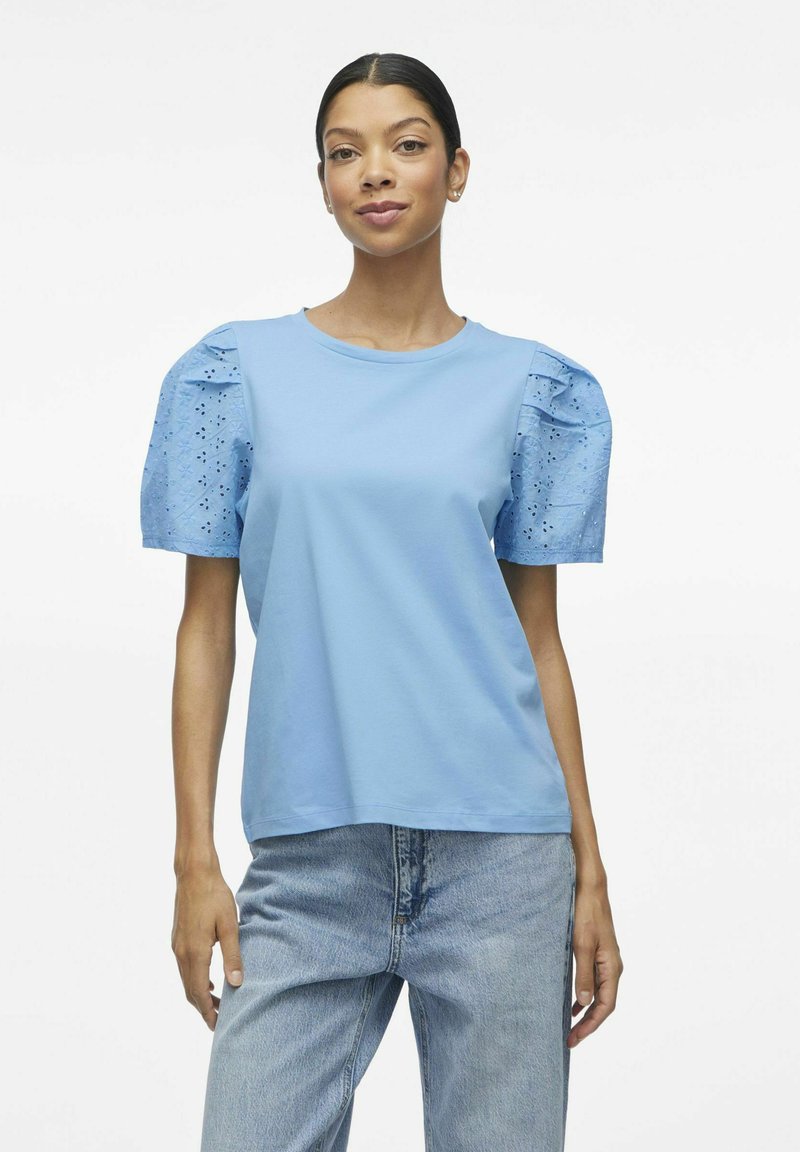 Jasnoniebieska bawełniana koszulka z krótkimi bufkami, z detalami w formie ażurowych wstawek. Prosty okrągły dekolt, luźny krój i gładka tekstura.