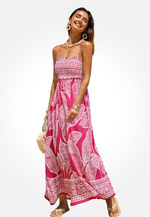 Vrouw glimlachend en poserend in een maxi-jurk met roze-wit patroon en spaghetti bandjes, met een geweven handtas, sandalen en grote oorbellen.