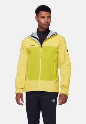 Uomo che indossa una giacca da esterno Mammut bicolore gialla con tasche con cerniera e pantaloni neri su uno sfondo semplice.