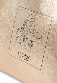 Roségouden pouch met reliëfstructuur; bevat een zwart lijngetekend gezicht en tropische bladeren, samen met de tekst "nōbo EST MMXV."