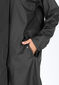 Black waterproof coat made of synthetic material, featuring snap button closures and side pockets for convenience. Smooth texture.