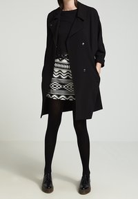 Cappotto nero doppiopetto con collo a revers, abbinato a una mini gonna a fantasia con design geometrici in bianco e nero.