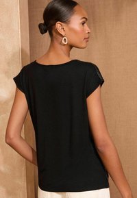 Juodas trumpų rankovių viršus su laisvu siluetu ir tekstūruotais pečių akcentais. Minkšta medžiaga ir apvalus iškirpė, rodomas iš nugaros.