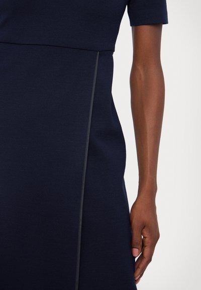 Robe de marine à manches courtes, en tissu texturé, avec un détail de couture latérale grise contrastante. La vue rapprochée montre une main reposant sur le tissu.