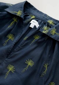 Navyblauwe blouse met een kraag, voorzien van groene bloemenborduursels, zachte stof en een lichte plooidetail onder de halslijn. Merkamet is zichtbaar.