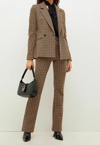 Brauner gemusterter Anzug mit einem doppelt geknöpften Blazer und schlank geschnittenen Hosen. Kombiniert mit einem schwarzen Oberteil und einer Handtasche. Glänzende schwarze High Heels.