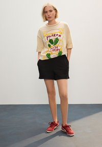 Cremefarvet grafisk t-shirt med design af grøn peber, kombineret med sorte shorts og røde sneakers. Bomuldsmateriale, løs pasform, korte ærmer.