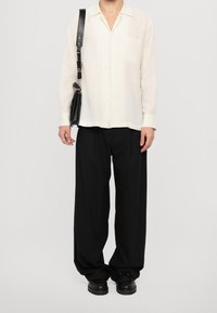 Camicia button-up in lino beige chiaro con tasca, abbinata a pantaloni neri larghi e scarpe nere. Include una borsa a spalla nera.