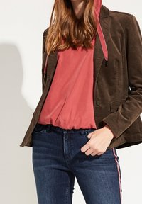 Ruskea corduroy-takki, jonka alla punainen huppari, yhdistetty tummansinisiin farkkuihin, joissa on sivulla vaaleanpunainen raita.