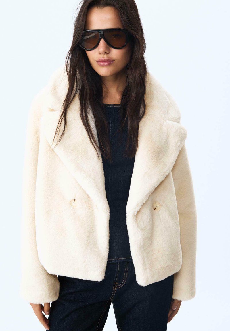 Creme faux fur jakke med sjalskrave, dobbeltrækket design og to knapper, parret med en mørk top og denim.