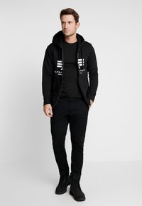 Schwarze Zip-up Kapuzenjacke mit weißem Logo, getragen über einem eng anliegenden schwarzen T-Shirt. Dazu schwarze Hosen und schwarze Schnürstiefel. Schlichtes Design.