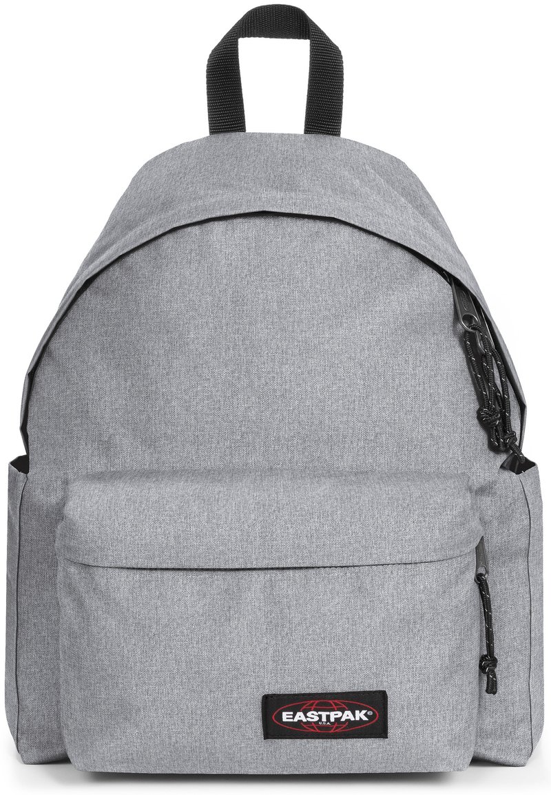 Mochila de tela gris con parte superior redondeada, bolsillo frontal, herrajes negros y un asa superior. Presenta una superficie texturizada y un parche con el logo.