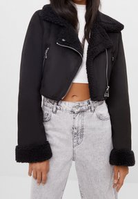 Avskuren svart jacka med fårskinnskrage och ärmslut, dragkedja framtill och två sidofickor; matchas med högmidjade grå jeans.