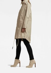 Beżowa lekka kurtka z wysokim kołnierzem, dwoma bocznymi kieszeniami i regulowanym ściągaczem u dołu, w zestawie z czarnymi obcisłymi dżinsami i beżowymi botkami za kostkę.