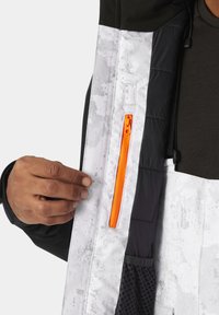 Innenseite einer weiß-schwarzen Jacke mit einer orangefarbenen Reißverschlusstasche, gestepptem Material und matter Oberfläche mit leichtem Steinmuster.
