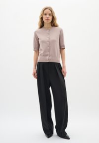 Hellrosa Kurzarm-Cardigan mit Knöpfen, weicher Struktur, runder Ausschnitt, kombiniert mit schwarzen weiten Hosen und spitzen schwarzen Schuhen.