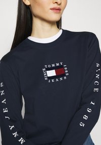 Lahetti pitkähihaine T-paita tummansinisestä puuvillasta, jossa on valkoinen pyöreä pääntie. Rintataskussa on logo, jossa on punaisia, valkoisia ja sinisiä korostuksia ja tekstiä hihojen varrella.