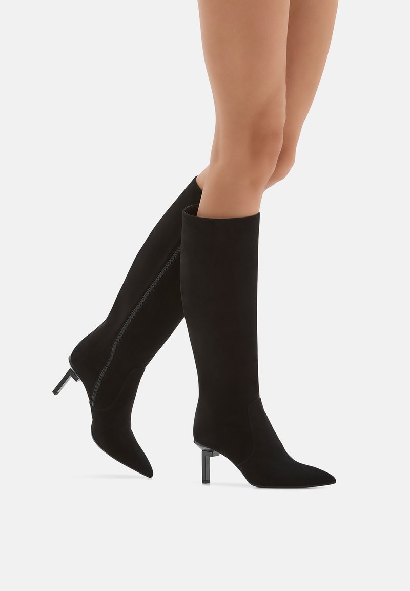 Gambe che indossano eleganti stivali neri a ginocchio con punte appuntite e tacchi bassi, in piedi su uno sfondo bianco neutro.