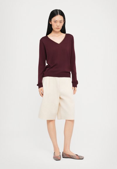 Bordeauxfarbene V-Ausschnitt-Pullover, cremefarbene, gefaltete Shorts und mehrfarbige, gestreifte Ballerinas. Der Pullover hat lange Ärmel und einen taillierten Saum.