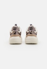 Rear view of sneakers with white fabric, brown accents, and a shiny dark brown heel. Features lace loops and "SM" embossed on the heel.