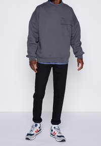 Grauer Sweatshirt mit Rundhalsausschnitt und einer Fronttasche, kombiniert mit schwarzen Hosen und grauen Sneakern mit blauen und roten Akzenten.