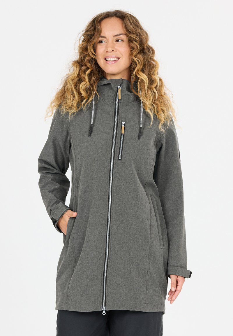 Femme aux cheveux bouclés portant une longue veste grise à fermeture éclair avec un col haut et les mains dans les poches, souriante et regardant vers la gauche.