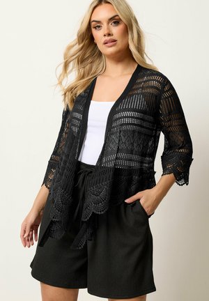 Kvinde med langt blond hår iført en sort åben strikket cardigan over en hvid top og sorte shorts, poserer med den ene hånd i lommen.