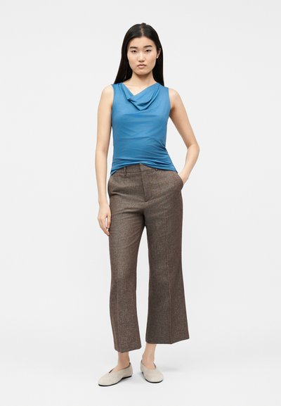 Γυναίκα σε όρθια στάση με το ένα χέρι στην τσέπη, φοράει μπλε αμάνικη μπλούζα με draping, καφέ cropped παντελόνι και μπεζ παπούτσια slip-on, πάνω σε απλό φόντο.