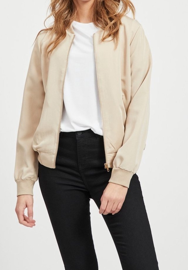 Femme portant une veste beige zippée sur une chemise blanche et un pantalon noir, debout avec une main détendue et l'autre légèrement pliée.