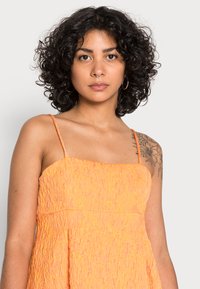 Vestido laranja texturizado com alças finas e decote quadrado. O tecido apresenta um design franzido e uma silhueta fluida.