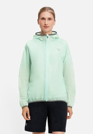 Frau trägt eine hellgrüne Kapuzenjacke mit Reißverschluss und schwarze Shorts, steht vor einem schlichten weißen Hintergrund.