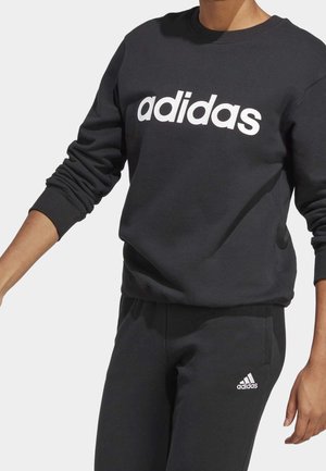 Person wearing black Adidas sweatshirt with large white logo and matching black sweatpants with small white Adidas logo.