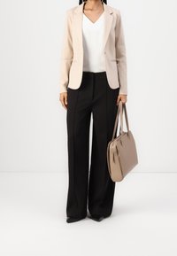 Beige kavaj, vit v-ringad topp, svarta vida byxor, svarta spetsiga skor, håller en beige handväska med detaljer i guld.