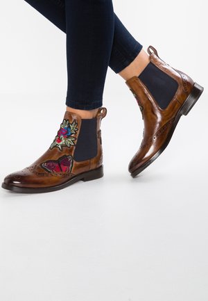 Bottes Chelsea en cuir marron avec panneaux latéraux élastiques, motifs brodés de fleurs et papillons, portées avec un pantalon sombre ajusté.