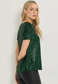 Top verde con paillettes, maniche corte, scollo rotondo e vestibilità ampia. Il tessuto ha una texture lucida e brilla alla luce.