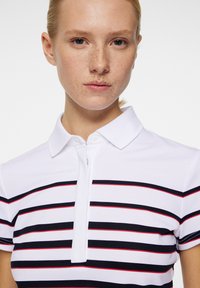 Vit polo skjorta med horisontella marinblå och röda ränder, med krage och halv dragkedja. Slät textur och åtsittande design.