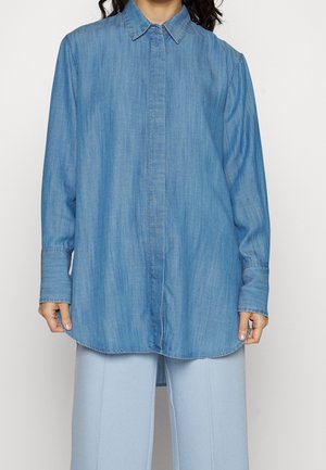 Woman wearing oversized light blue denim shirt with long sleeves and hidden button placket, paired with light blue wide-leg pants.