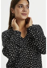 Schwarze Bluse mit weißen Punkten, V-Ausschnitt, langen Puffärmeln, leicht strukturierter Oberfläche und knöpfbaren Manschetten.