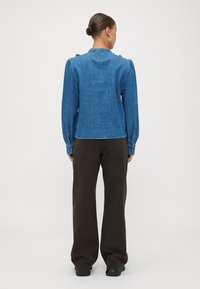 Jeanskjorte med lange ermer i blå farge, knappdetaljer på mansjettene og høy krage; kombinert med mørkebrune, løstsittende bukser.