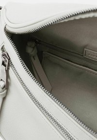 Interno di una borsa a tracolla bianca in pelle con rivestimento in tessuto grigio, chiusura con cerniera e tasca interna. Presenta un dettaglio del manico intrecciato.