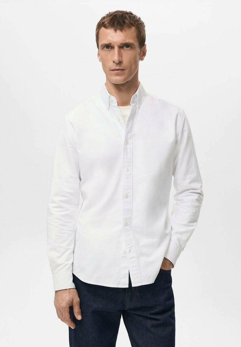 Camisa blanca de algodón con botones, con cuello, mangas largas y corte recto, presentada junto a pantalones vaqueros oscuros.