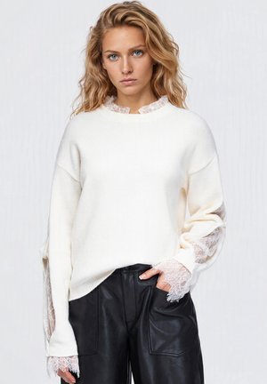 Une femme porte un pull blanc à col et poignets en dentelle, associé à un pantalon en cuir noir, sur un fond clair et uni.