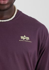 Chemise à manches longues violet en tissu doux, avec un petit logo doré à trois rayures et le texte "ALPHA INDUSTRIES" sur la poitrine.