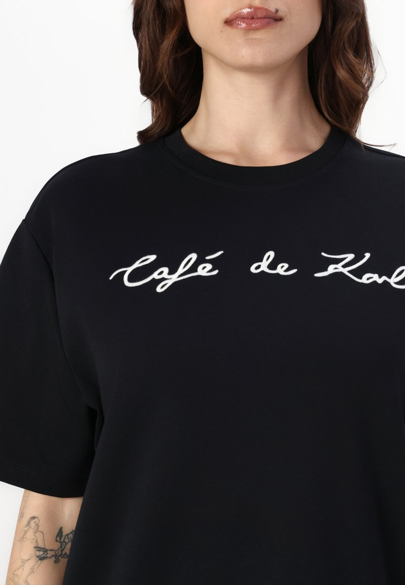 Schwarzes Baumwoll-T-Shirt mit weißem gesticktem Text, der auf der Vorderseite "café de K" lautet. Lockere Passform mit kurzen Ärmeln und rundem Ausschnitt.